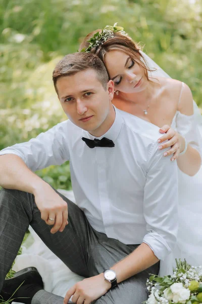 ドレスの美しいかわいい花嫁は緑の草の上に座っている白いシャツの後ろにスタイリッシュなグロムを抱きしめます 結婚式の肖像画 恋の新婚旅行の写真 — ストック写真