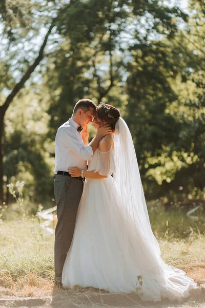 Fashionable groom and cute bride in white dress with tiara of fresh flowers, hugging, laughing in park, garden, forest outdoors. Wedding photography, portrait of smiling newlyweds.