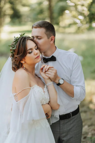 Fashionable groom and cute bride in white dress with tiara of fresh flowers, hugging, laughing in park, garden, forest outdoors. Wedding photography, portrait of smiling newlyweds.