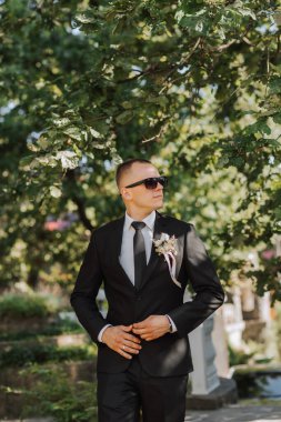 Kafasında siyah gözlük, beyaz gömlek ve kravat olan, klasik siyah takım elbiseli şık bir damadın açık hava portresi.