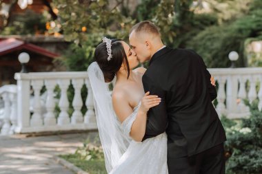 Bahçede genç ve güzel bir düğün çifti portresi omuzları açık gelinlik içinde gelin, siyah klasik takım elbiseli damat doğanın arka planında şefkatle öpüşüyor.