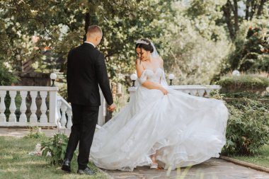 Bahçede genç ve güzel bir düğün çifti portresi, omuzları açık gelinlik içinde gelin daire çiziyor ve elbisesiyle hava atıyor, damat siyah klasik takım elbise giymiş.