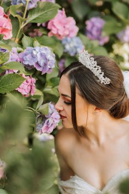 Gelinlik giymiş, omuzları açık, başında taçla ortanca çiçeklerinin yanında oturan genç ve güzel bir gelin. Sert güneş ışığı altında çekilmiş bir moda fotoğrafı.