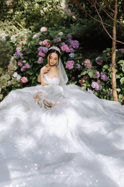 Gelinlik giymiş, omuzları açık, başında taçla ortanca çiçeklerinin yanında oturan genç ve güzel bir gelin. Sert güneş ışığı altında çekilmiş bir moda fotoğrafı.