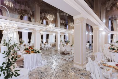 Düğün salonunun içi beyaz renklerde, masalar hazır, her şey çiçeklerle süslenmiş, kristal avizeler asılmış, tavandaki yansıma ses katıyor.