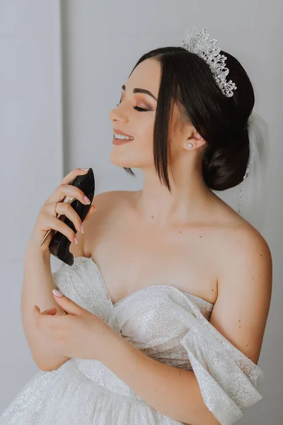 a brunette bride in a chic dress, hairdo and a tiara on her head sprays perfume on her body in her room