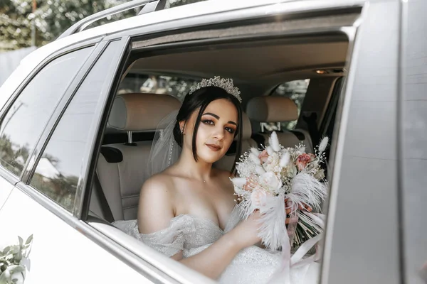 the bride sits in a white luxury car on the wedding day with a bouquet. Portrait of the bride. lush white lace dress.