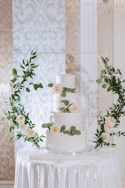 a four-tiered white wedding cake decorated with white roses stands on a table at the entrance to the wedding hall