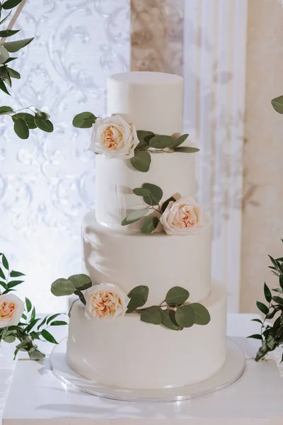 a four-tiered white wedding cake decorated with white roses stands on a table at the entrance to the wedding hall