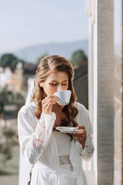 Beyaz bornozlu güzel bir gelin büyük bir şehre tepeden bakan bir otel balkonundaki beyaz bardaktan kahve içer. Evlenmeden önceki gün.