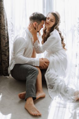 Beyaz cübbeli güzel, seksi gelin, beyaz gömlekli damat gelini kucaklıyor ve otel odasındaki bir pencerenin arkasından öpüyor. Yeni evli çiftlerin düğün portresi. Dikey fotoğraf.