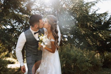 Genç bir düğün çifti, parkta bir yaz ormanının arka planında romantik anlar yaşamaktan zevk alır. Beyaz gelinlikli gelin, beyaz gömlekli damat, yelek ve papyonlu gelin sarıl ve öp gelini.