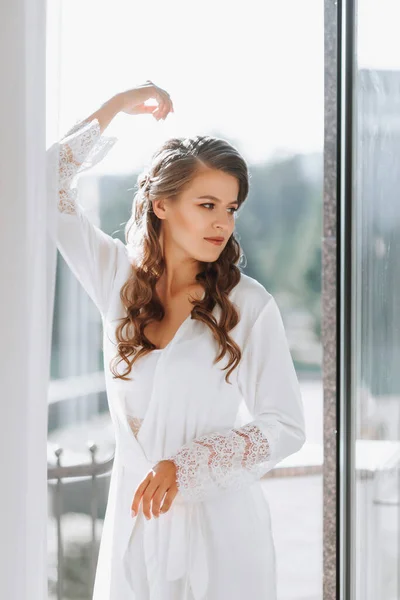 A beautiful bride in a white robe drinks coffee from a white cup on a hotel balcony overlooking a big city. On your wedding day before marriage.