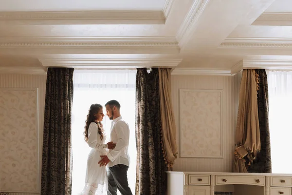 Beautiful, sexy bride in a white robe, groom in a white shirt hugs the bride in a hotel room. Wedding portrait of newlyweds in love. Free space.