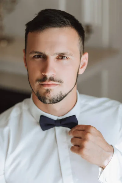 A stylish groom with a beard is preparing for the wedding ceremony. Groom\'s morning. A businessman fastens a black bow tie. The groom is getting ready in the morning before the wedding ceremony