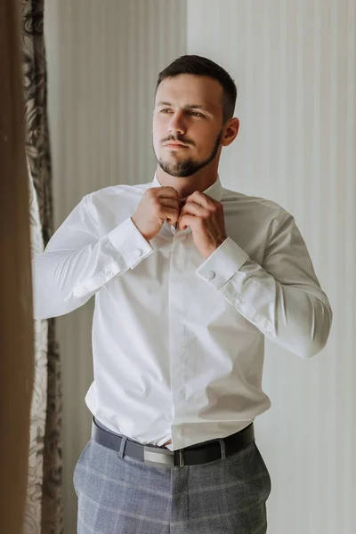 A stylish groom with a beard is preparing for the wedding ceremony. Groom\'s morning. A businessman is buttoning up a white shirt. The groom is getting ready in the morning before the wedding ceremony