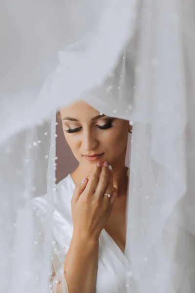A luxurious bride with a classic hairstyle, wearing a long robe and a long veil, stands by the window under a veil in her room. The bride on a white background
