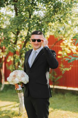 Damadın klasik siyah takım elbiseli ve gözlüklü fotoğrafı, elinde bir buket çiçekle.