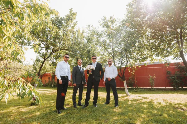 The groom in a black suit and glasses and his stylish friends wearing white shirts and black pants and glasses are standing in the backyard in the garden. The groom is holding a bouquet.