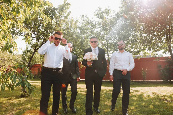 The groom in a black suit and glasses and his stylish friends wearing white shirts and black pants and glasses are standing in the backyard in the garden. The groom is holding a bouquet.