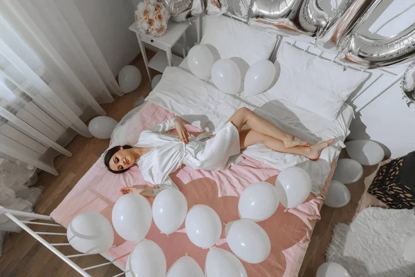 A brunette bride in a white robe and veil lies on her bed near white balls. Wedding concept. Morning of the bride