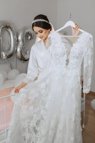 白いペチコートの美しい若い花嫁がベッドのそばに立って 彼女のウェディングドレスに触れています 結婚式のための最終準備 花嫁は花嫁を待っている 朝から花嫁 — ストック写真