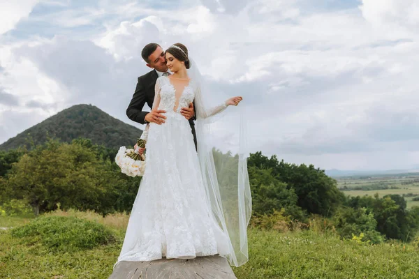 stylish bride and groom hug and kiss against the background of summer mountains. the concept of a rustic wedding in the mountains, happy bohemian newlyweds.