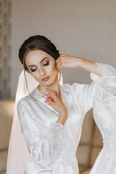 A beautiful woman with hair on her head, make-up and a wedding veil in a dressing gown is standing next to her dress on a mannequin. Dressing up and preparing for the wedding ceremony