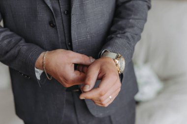 Pencerenin yanındaki bir odada, klasik gri takım elbiseli damadın portresi. Adamın biri altın rengi bir kol saati takıyor, yakın plan.