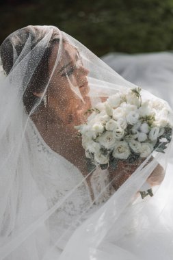 Gelinlik içinde uzun bir tren ve duvağı olan bir gelin yeşil çimlerin üzerinde bir duvağın altında otururken bir gül buketi tutuyor. Yukarıdan Fotoğraf