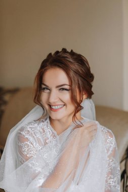 Gelinin beyaz bornozlu ve zarif saç stili olan sabahı. Yetişkin bir kız sandalyeye oturur ve bir küpe takar, kameranın lensine bakar ve gülümser.