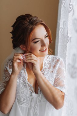 Gelinin beyaz bornozlu ve zarif saç stili olan sabahı. Yetişkin bir kız sandalyeye oturur ve küpesini takar, pencereden dışarı bakar ve gülümser. Kahverengi saçlı