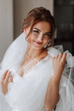 Sabahlık giymiş kahverengi saçlı bir gelin kameraya poz veriyor, gelinliğine bakıyor. Şık saç stili. Güzel makyaj. Gönüllü duvak. Gelinin sabahı