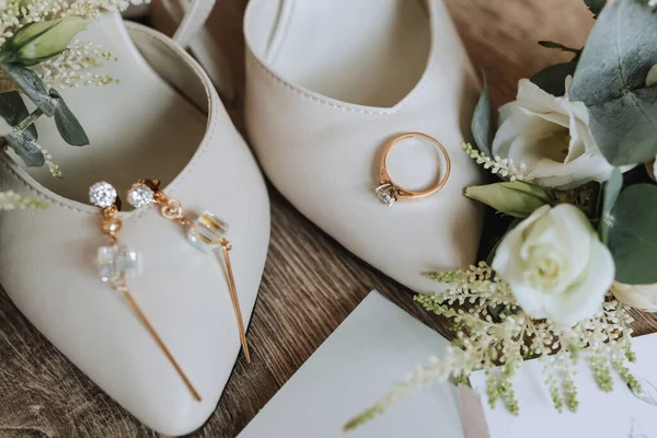 White wedding shoes and details of the bride. Wedding ring with diamond and gold bracelet with crystals, on wooden background. Flowers and greenery. Wedding theme