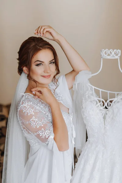 A bride with brown hair in a robe, posing for the camera, looking at her wedding dress. Elegant hairstyle. Nice makeup. Voluminous veil. Morning of the bride