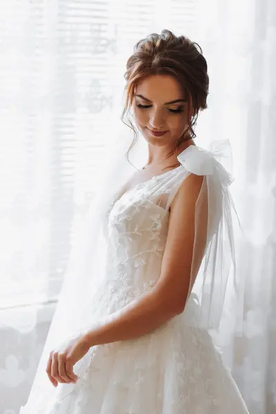 A bride with brown hair in an elegant white dress poses for the camera. Elegant hairstyle. Nice makeup. Voluminous veil. Morning of the bride
