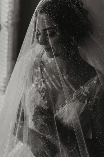 The bride is dressed in an elegant dress, covered with a veil, posing and holding a boutonniere. Wedding black and white photo, morning of the bride