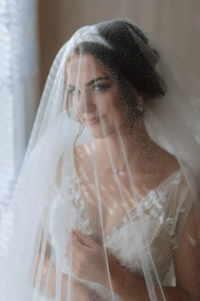 The bride is dressed in an elegant dress, covered with a veil, posing and holding a boutonniere. Wedding photo, morning of the bride