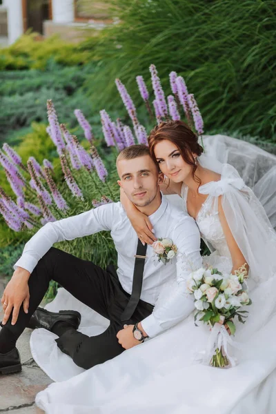 Portrait of the bride and groom in the park. The bride is holding a bouquet and hugging her groom, they are sitting, posing near lavender flowers. Wedding walk in the park. Long train of the dress