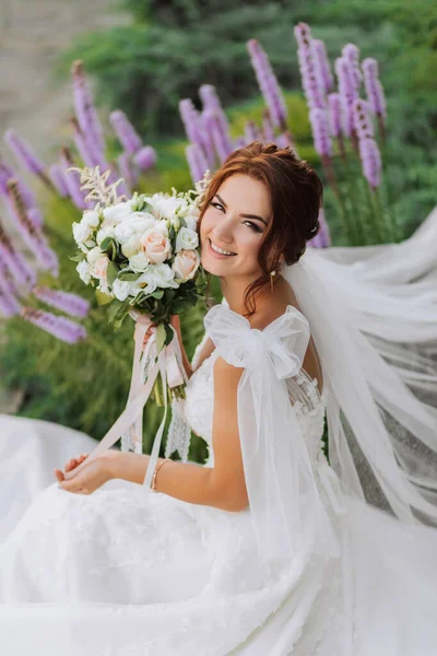 公園内の花嫁の肖像画 エレガントなドレスの花嫁は ラベンダーの花の近くにポーズする花束を保持しています 結婚式は公園で散歩する ドレスのロングトレイン — ストック写真