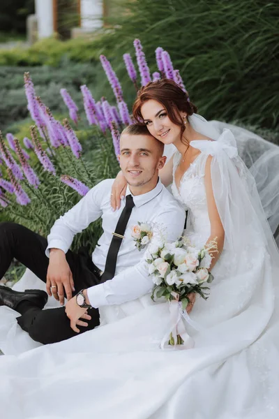 Portrait of the bride and groom in the park. The bride is holding a bouquet and hugging her groom, they are sitting, posing near lavender flowers. Wedding walk in the park. Long train of the dress