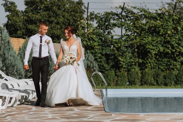 Portrait of the bride and groom. The bride and groom walk by the pool holding hands. Wedding walk in the park. Long train of the dress