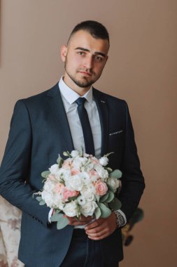 Koyu mavi takım elbiseli, elinde bir buket çiçekle kendine güvenen zarif, yakışıklı bir adam. Damat düğün töreni için hazırlanıyor.