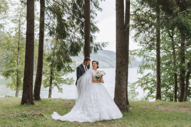 Bir düğün çifti, hayatlarının en güzel gününü bir gölün ve uzun ağaçların önünde geçiriyor. Damat geline arkadan sarılıyor.