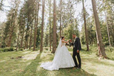 Modaya uygun bir damat ve beyaz elbiseli şirin bir gelin tren ve kafasında taçla parkta, bahçede, ormanda mutlu bir şekilde yürüyor. Düğün fotoğrafı, gülümseyen yeni evlilerin portresi..