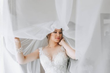 Gelinin portresi. Gelin duvaklı, yaka çiçeğiyle beyaz bir arka planda poz veriyor. Güzel saç ve makyaj. Elbise için açık büst. Kristalli diyadem