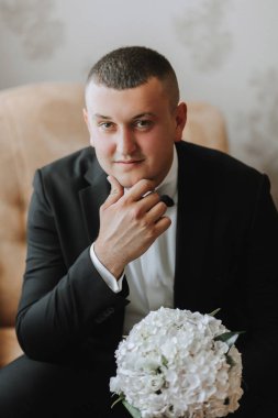 Portre. Beyaz gömlekli, siyah takım elbiseli bir adam koltukta oturuyor, elinde buketle poz veriyor. Şık bir saat. Erkek tarzı. Moda. İş dünyası