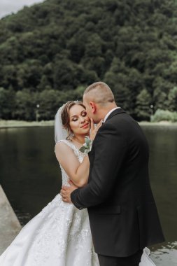 Uzun elbiseli esmer bir gelin ve klasik takım elbiseli bir damat arka planda şatosu olan gölün yakınındaki bir köprüde birbirlerine sarılıyorlar..