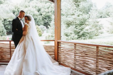 Düğün portresi. Şık elbiseli gelin ve klasik takım elbiseli damat çardakta birbirlerine sarılıyorlar. Uzun tren. Yaz düğünü