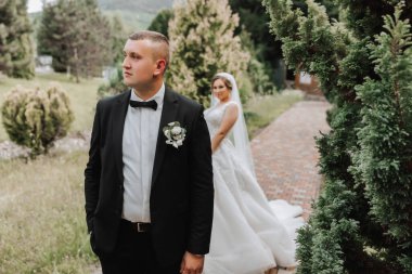Düğün portresi. Damat ön planda duruyor. Gelin zarif bir elbise içinde, damat ise yeşil ağaçların arka planına karşı klasik bir takım elbise içinde. Nazikçe dokun. Yaz düğünü. Doğada bir yürüyüş